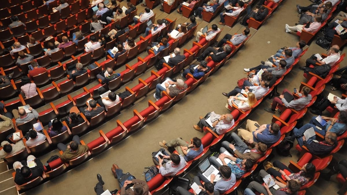 Konferans salonunda yukarıdan görünen, dizüstü bilgisayar ve belgelerle geniş bir salonu dolduran katılımcılar