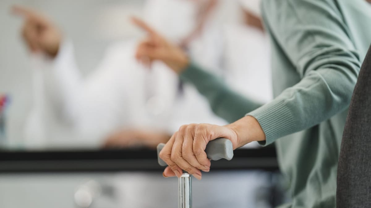 Klinikte baston tutan bir kişinin eli, arka planda beyaz önlüklü bir hekim